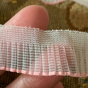 May include: White and pink ruffled trim with a delicate, grid-like pattern. The trim has a pink edge.