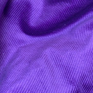 Pode incluir: Close-up de um tecido roxo vibrante com textura de veludo cotelê. O tecido tem uma aparência macia e aveludada com reflexos de luz sutis, criando um visual rico e luxuoso. O padrão canelado adiciona profundidade e interesse visual ao têxtil.