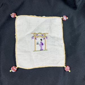 Peut inclure: Carré de tissu blanc brodé avec une bordure dorée et des coins perlés. La broderie représente une figure en kimono violet sous une porte japonaise traditionnelle. La porte a des accents violets et verts.