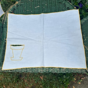 May include: White linen napkin with a yellow embroidered flower pot design. The napkin has a yellow border and is folded. The napkin is on a green wicker chair.