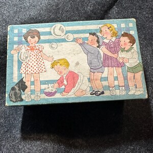 May include: A vintage box with a blue and white striped background featuring a group of children playing with soap bubbles. The children are dressed in colorful clothing and there is a black cat in the lower left corner of the box.