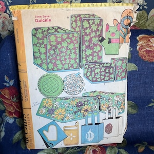May include: A vintage McCall's Time Saver Quickie sewing pattern envelope featuring illustrations of kitchen appliance covers, a pig-shaped oven mitt, tote bags, and circular hot pads with floral and clover patterns, topped with a yellow measuring tape.