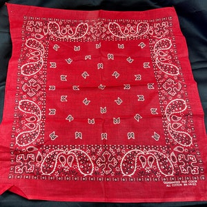 May include: A red bandana with a white and black paisley pattern. The bandana has the text "GUARANTEED FAST COLOR ALL COTTON IN 14193" printed on the bottom.