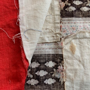 May include: A close-up of a patchwork fabric with red, white, and brown sections. The fabric is made of different textures and patterns, including a floral print and a geometric design. The fabric is sewn together with visible stitching.