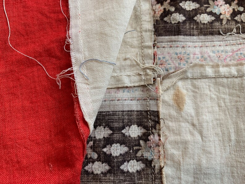 May include: A close-up of a patchwork fabric with red, white, and brown sections. The fabric is made of different textures and patterns, including a floral print and a geometric design. The fabric is sewn together with visible stitching.