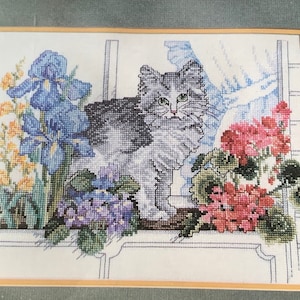 Puede incluir: Un patrón de punto de cruz de un gato gris sentado en una ventana con iris azules, flores moradas y geranios rojos.