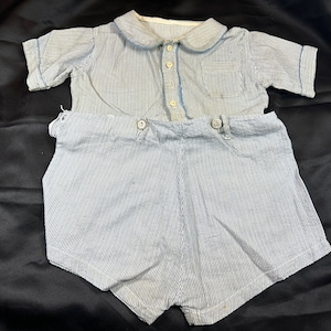 May include: A blue and white striped short-sleeved shirt and matching shorts set for a baby or toddler. The shirt has a white collar and a button-up front. The shorts have a button closure and a waistband.