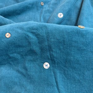 Könnte beinhalten: Nahaufnahme eines teal-farbenen Cordstoffs mit mehreren Knöpfen. Der Stoff hat eine gerippte Textur und ist einfarbig teal. Die Knöpfe sind rund und haben einen perlmuttartigen Glanz.