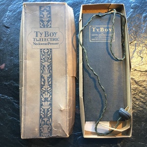 Puede incluir: Una caja vintage para un planchador eléctrico de corbatas llamado "Ty Boy The Electric Neckwear Presser". La caja es marrón con un diseño floral decorativo y la tapa está abierta para revelar un cable y un enchufe negros.