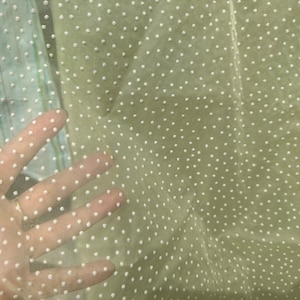 May include: A sheer, light beige fabric with a white polka dot pattern. The fabric is translucent and has a soft, delicate texture.