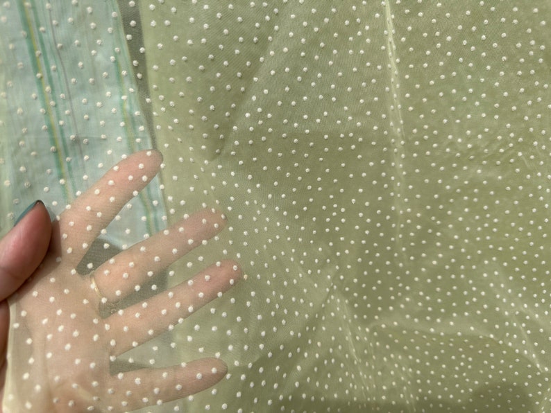 May include: A sheer, light beige fabric with a white polka dot pattern. The fabric is translucent and has a soft, delicate texture.