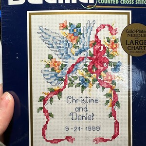May include: A counted cross stitch kit featuring a white dove with red ribbon and flowers. The kit includes a gold-plated needle and a large chart. The kit is for a wedding and includes the names Christine and Daniel and the date 5-21-1999.