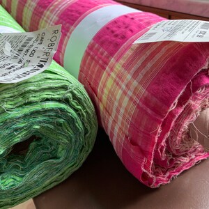 May include: Two rolls of fabric, one green and one pink with white and yellow plaid pattern. The green fabric is rolled tightly and the pink fabric is rolled loosely. Both rolls have price tags attached.