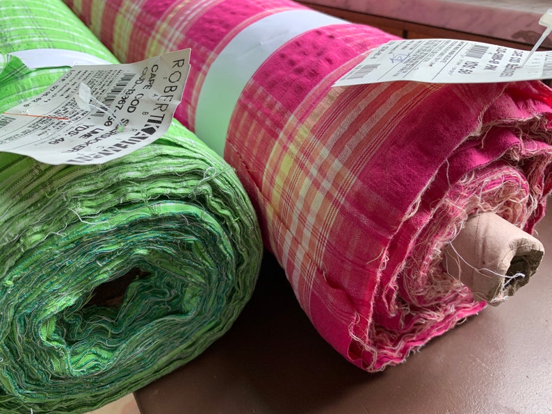 May include: Two rolls of fabric, one green and one pink with white and yellow plaid pattern. The green fabric is rolled tightly and the pink fabric is rolled loosely. Both rolls have price tags attached.