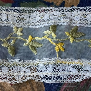 Könnte beinhalten: Weiße Spitzenborte mit einem zarten floralen Stickmuster in Grün- und Gelbtönen. Die Borte hat einen gewellten Rand und ein sich wiederholendes Muster aus Blättern und Blüten.