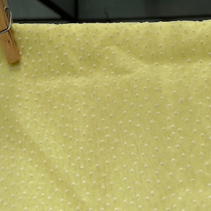 May include: A pale yellow fabric with a subtle white polka dot pattern. The fabric is sheer and has a slightly textured surface.