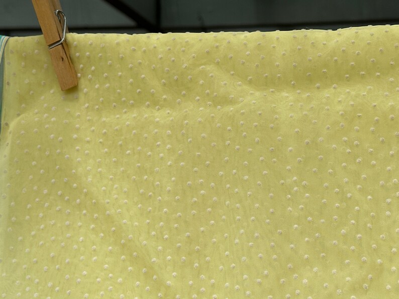 May include: A pale yellow fabric with a subtle white polka dot pattern. The fabric is sheer and has a slightly textured surface.