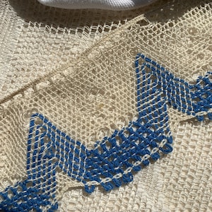 May include: Close-up of a white crocheted lace trim with a blue geometric pattern. The lace is delicate and intricate, with a scalloped edge.