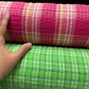 May include: Two rolls of fabric with a plaid pattern. The top roll is pink with white and yellow stripes. The bottom roll is green with white stripes.