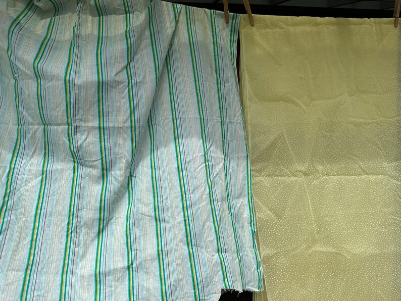 May include: Two pieces of fabric hanging on a clothesline. One piece is a light blue fabric with green, white, and purple stripes. The other piece is a light yellow fabric with white polka dots.