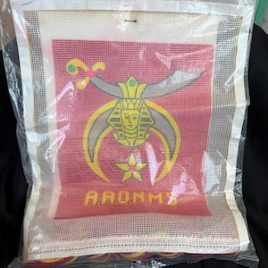 Puede incluir: Lienzo de punto de aguja con fondo rojo y emblema amarillo y gris. El emblema incluye una cabeza de faraón, una estrella y la palabra "AARONMA" en amarillo. El lienzo está empaquetado en plástico transparente.