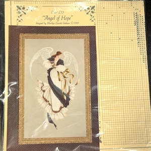 Puede incluir: Un kit de punto de cruz titulado "Angel of Hope", diseñado por Marilyn Leavitt-Imblum en 1991. La imagen presenta un punto de cruz enmarcado de un ángel con un vestido fluido con alas, detalles dorados y una faja azul oscuro. El kit incluye un gráfico.