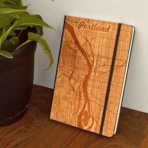 May include: A wooden notebook with a laser-etched map of Portland, Oregon, featuring the city's layout and the word "Portland" at the top. The notebook has a black elastic band and is set on a wooden surface.