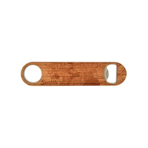 May include: Wooden bottle opener with a map of Seattle etched into the surface. The opener has a circular hole on one end and a bottle cap opener on the other. The word "Seattle" is engraved on the wood.