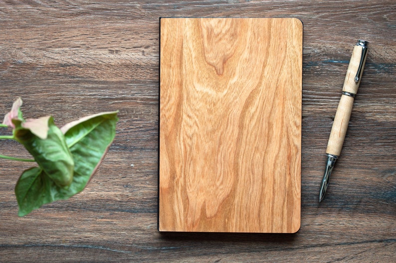 May include: A rectangular notebook with a light brown wood grain cover and a matching light brown wood grain pen with silver metal accents. The notebook is positioned vertically, and the pen is placed to the right of the notebook.