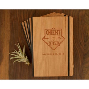 May include: A stack of light brown wooden notebooks with black elastic bands. The top cover features a laser-engraved Cheez-It Bowl logo with a desert landscape graphic and the text "CHEEZ-IT BOWL" followed by the date "DECEMBER 27, 2019".