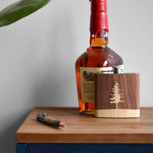 Pode incluir: Uma garrafa de madeira com um design de árvore cortada a laser, combinada com uma garrafa de líquido âmbar selada com cera vermelha. Uma caneta repousa sobre a superfície de madeira. A garrafa é feita de madeira escura com uma base de madeira clara.
