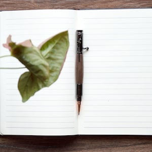 A black walnut bolt action pen lays in the spine of one of our white journals, sitting on top of a desk.