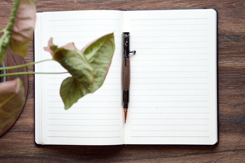 A black walnut bolt action pen lays in the spine of one of our white journals, sitting on top of a desk.