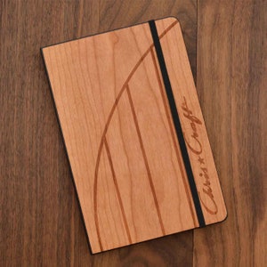 May include: A rectangular notebook with a light brown wood grain cover featuring a curved dark brown inlay design. A vertical black elastic band secures the cover. The text Chris-Craft is engraved on the right side of the wooden front cover.