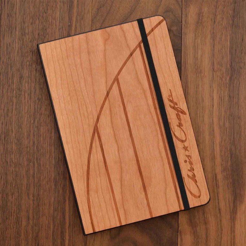 May include: A rectangular notebook with a light brown wood grain cover featuring a curved dark brown inlay design. A vertical black elastic band secures the cover. The text Chris-Craft is engraved on the right side of the wooden front cover.