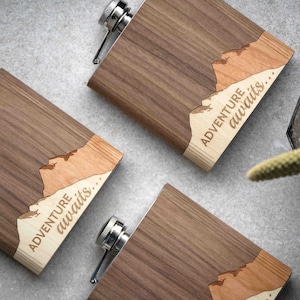 Puede incluir: Tres petacas de madera con tapón de acero inoxidable. Las petacas tienen un acabado de veta de madera oscura con un diseño de montaña de madera clara y la inscripción "ADVENTURE awaits..." grabada. Sobre una superficie gris.
