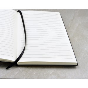 May include: A black lined notebook with a black ribbon bookmark. The notebook is open to a blank page.