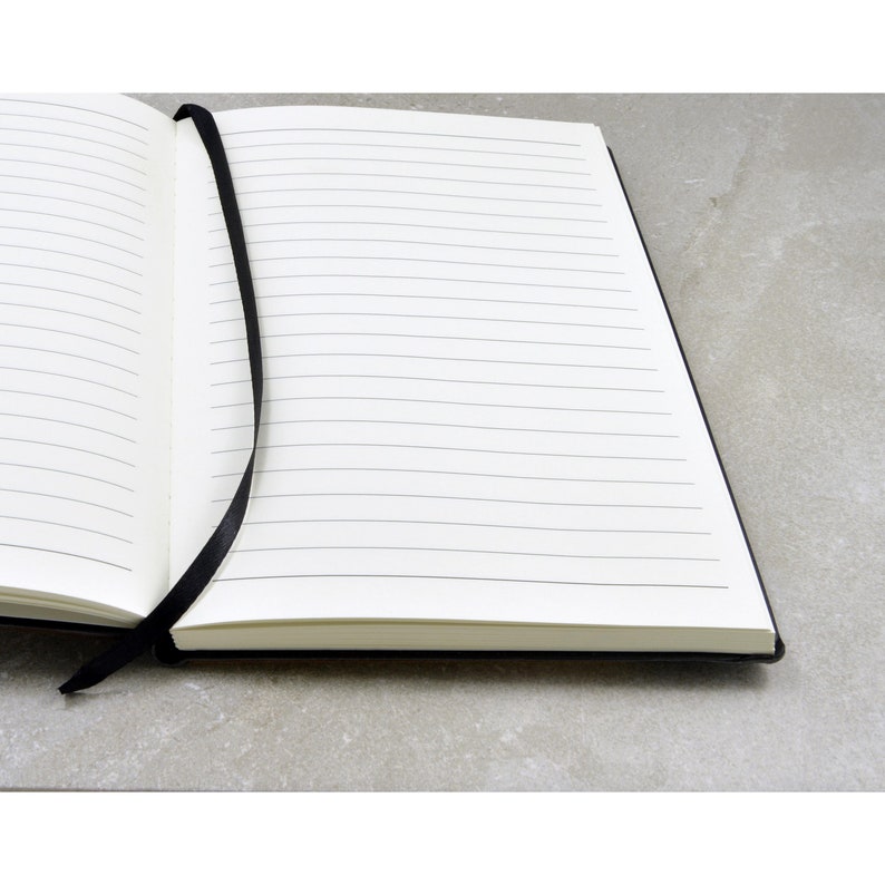 May include: A black lined notebook with a black ribbon bookmark. The notebook is open to a blank page.