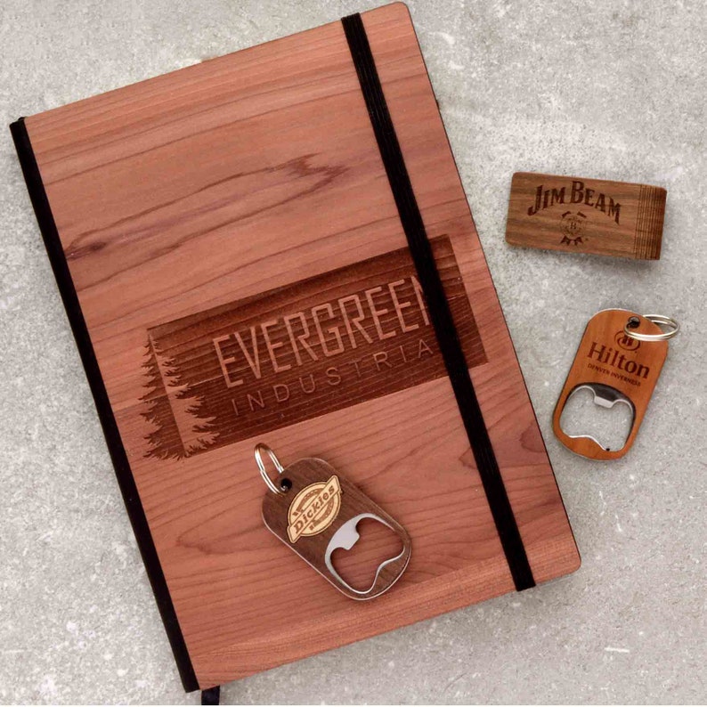 May include: A wooden journal with a black elastic closure and a laser-engraved logo that reads "EVERGREEN INDUSTRIES". The journal is accompanied by three wooden bottle openers with different logos: "Dickies", "Hilton Hotels", and "Jim Beam".