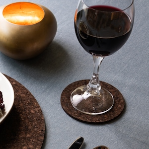 May include: A glass of red wine on a cork coaster. The glass is clear and has a long stem. The coaster is round and brown.