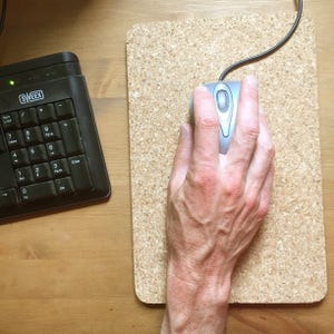 May include: A hand using a silver computer mouse on a cork mousepad.