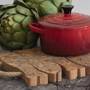 May include: Red cast iron pot with a black knob on the lid, sitting on a cork trivet with a rope handle. The pot has the words "Le Creuset" on the side.