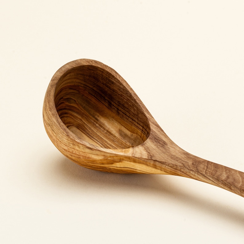 May include: A wooden olive wood ladle with a natural wood finish. The ladle has a long handle and a deep, rounded bowl.