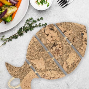 May include: A cork fish-shaped chopping board with grey detailing. The board has a handle with a rope loop. The cork has a natural, marbled pattern. A plate of roasted vegetables and a small bowl of sauce are in the background.