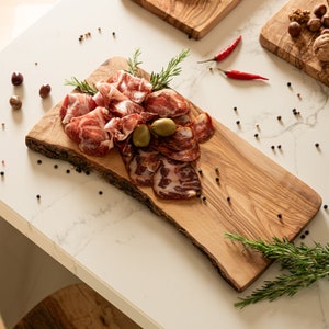 May include: A wooden serving board with a natural edge, featuring a selection of cured meats, olives, and rosemary sprigs. The board is on a white marble surface.