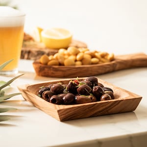 May include: A small wooden bowl filled with dark olives. The bowl is made of light brown wood and has a square shape. The olives are arranged in a pile in the centre of the bowl.