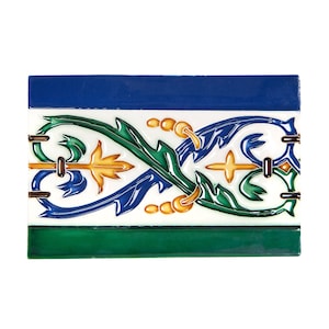May include: Rectangular ceramic tile with a decorative floral design. The tile features a white background with a border of blue and green. The floral design includes green, blue, and gold elements.