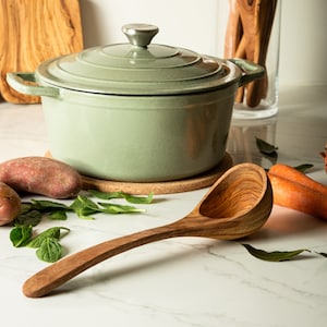 May include: A green cast iron pot with a lid and a wooden ladle. The pot is sitting on a cork trivet. There are potatoes and green leaves on the counter.