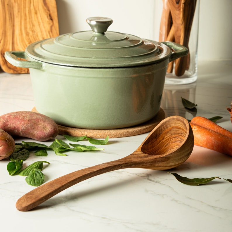 May include: A green cast iron pot with a lid and a wooden ladle. The pot is sitting on a cork trivet. There are potatoes and green leaves on the counter.