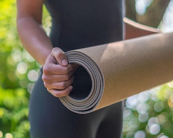Op de afbeelding: Opgerolde yogamat in bruin- en grijstinten. De mat wordt vastgehouden door een persoon die een zwarte outfit draagt, met een vage groene achtergrond. De mat lijkt van kurk te zijn.